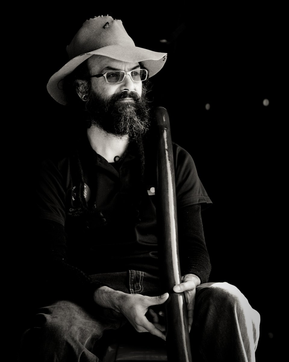 Listen — Black-and-white portrait of a bearded man in a wide-brimmed hat, seated and holding a didgeridoo