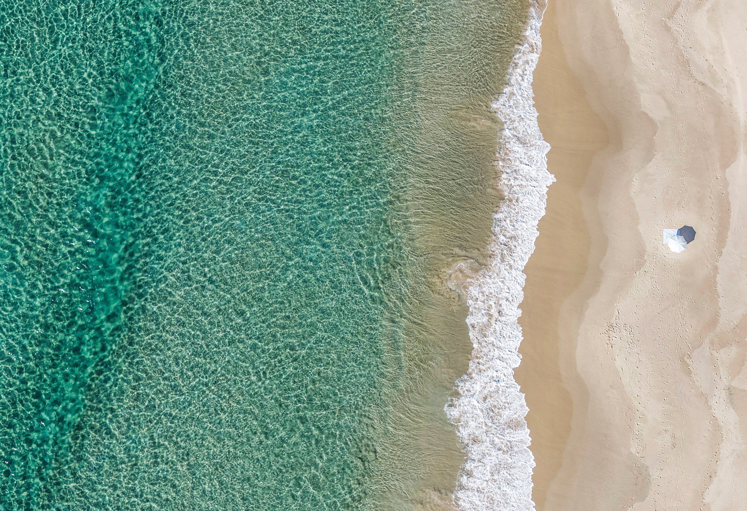 Sundays — Aerial view of a single beach umbrella on pale sand beside the shoreline and clear green water